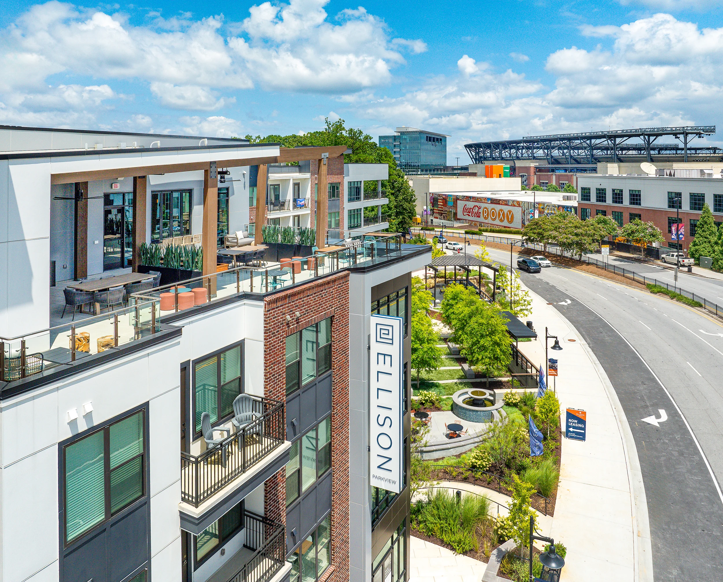 Modern Apartment Complex Overview Aerial view of a modern apartment complex with balconies, greenery, and a road leading to a stadium in the background under a sunny sky.
