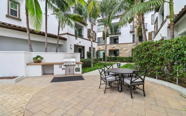 An outdoor patio area featuring a grill station, a round dining table with chairs, surrounded by greenery and palm trees.