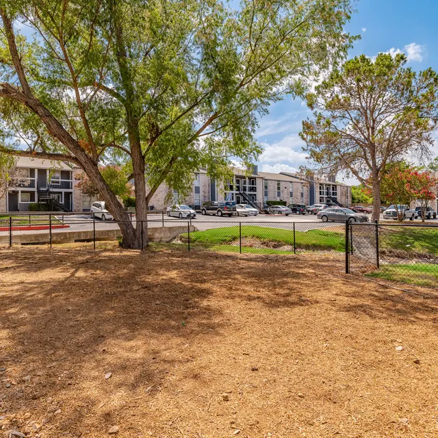A fenced green space featuring a large tree and benches, overlooked by apartment buildings under a clear blue sky.