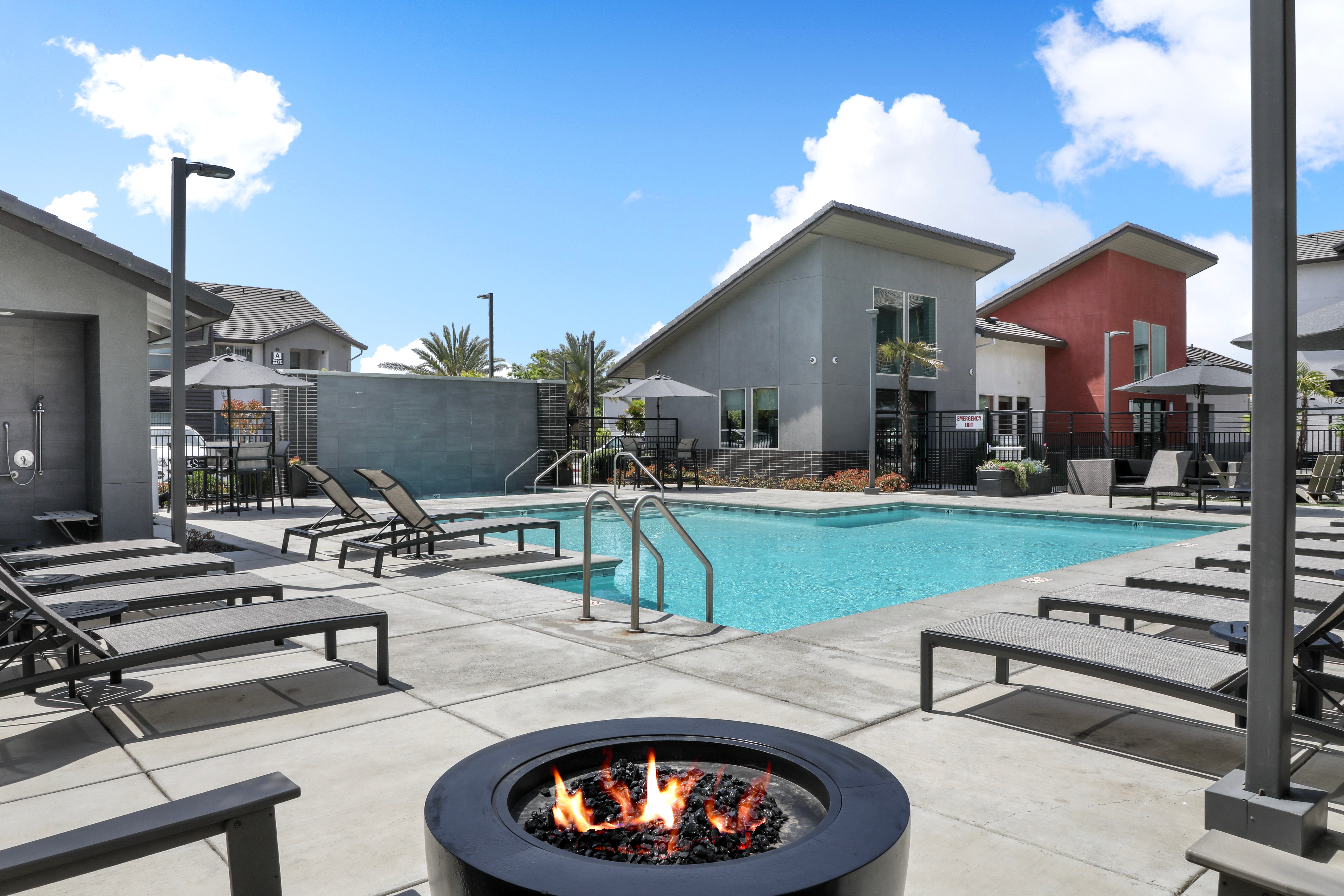 A swimming pool area featuring a fire pit, lounge chairs, and modern buildings in the background under a bright blue sky.