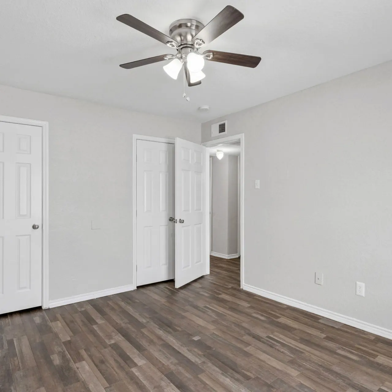 An empty room with wooden flooring, a ceiling fan, and light gray walls. Two closed doors are visible, leading to different areas.