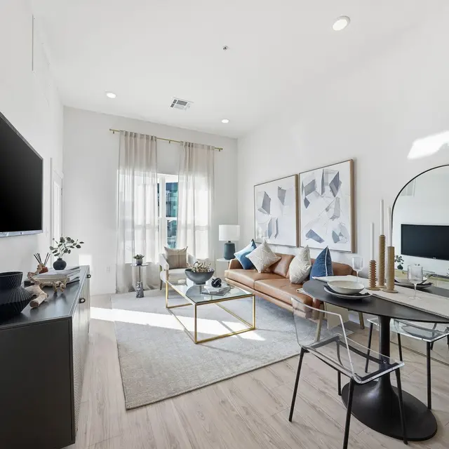 Modern living room with a sofa, coffee table, dining area, and large windows. Bright with neutral colors and stylish decor.