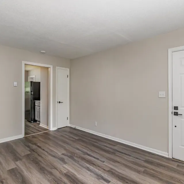 A spacious, empty room with light-colored walls and wooden flooring, featuring a closed door on the right and an open doorway leading to another room in the background.