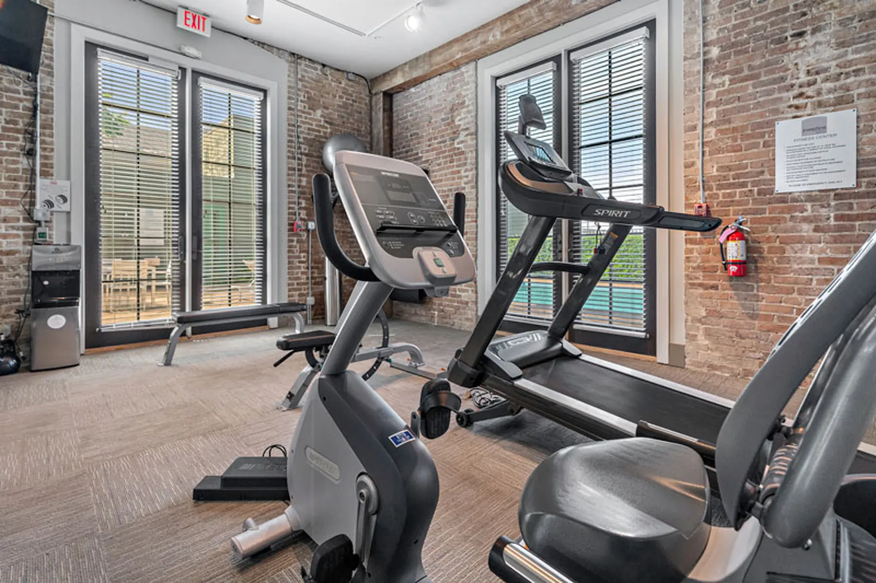 A gym area featuring exercise equipment with large windows and exposed brick walls.