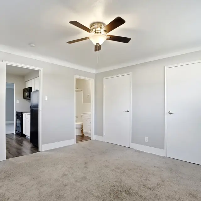 A spacious, modern bedroom featuring light gray walls, a ceiling fan, and multiple doors. The floor is covered with carpet, and there is a view into a bathroom and kitchen area through the open doors.