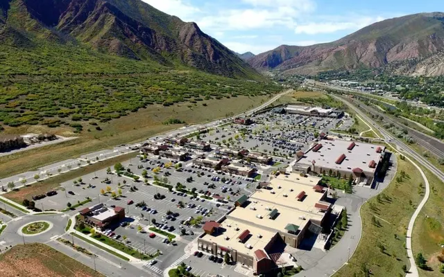 Aerial view of a large shopping center surrounded by mountains, featuring a spacious parking lot and retail buildings.