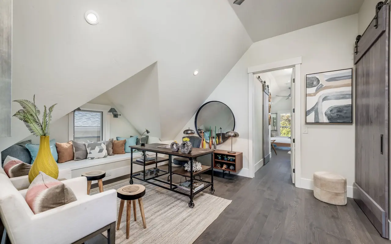 A cozy attic space featuring a stylish sofa with colorful cushions, a coffee table, and decor elements. The room has slanted ceilings, hardwood flooring, and bright lighting, creating a warm atmosphere.