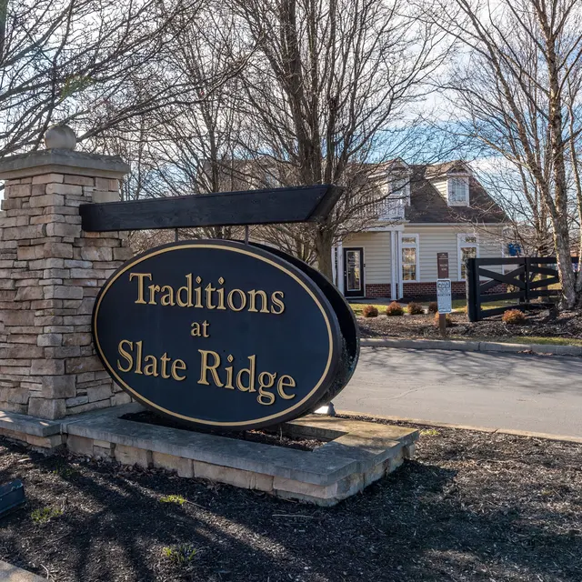 Sign for Traditions at Slate Ridge, a residential community or facility, situated along a paved road, surrounded by trees and a fence.