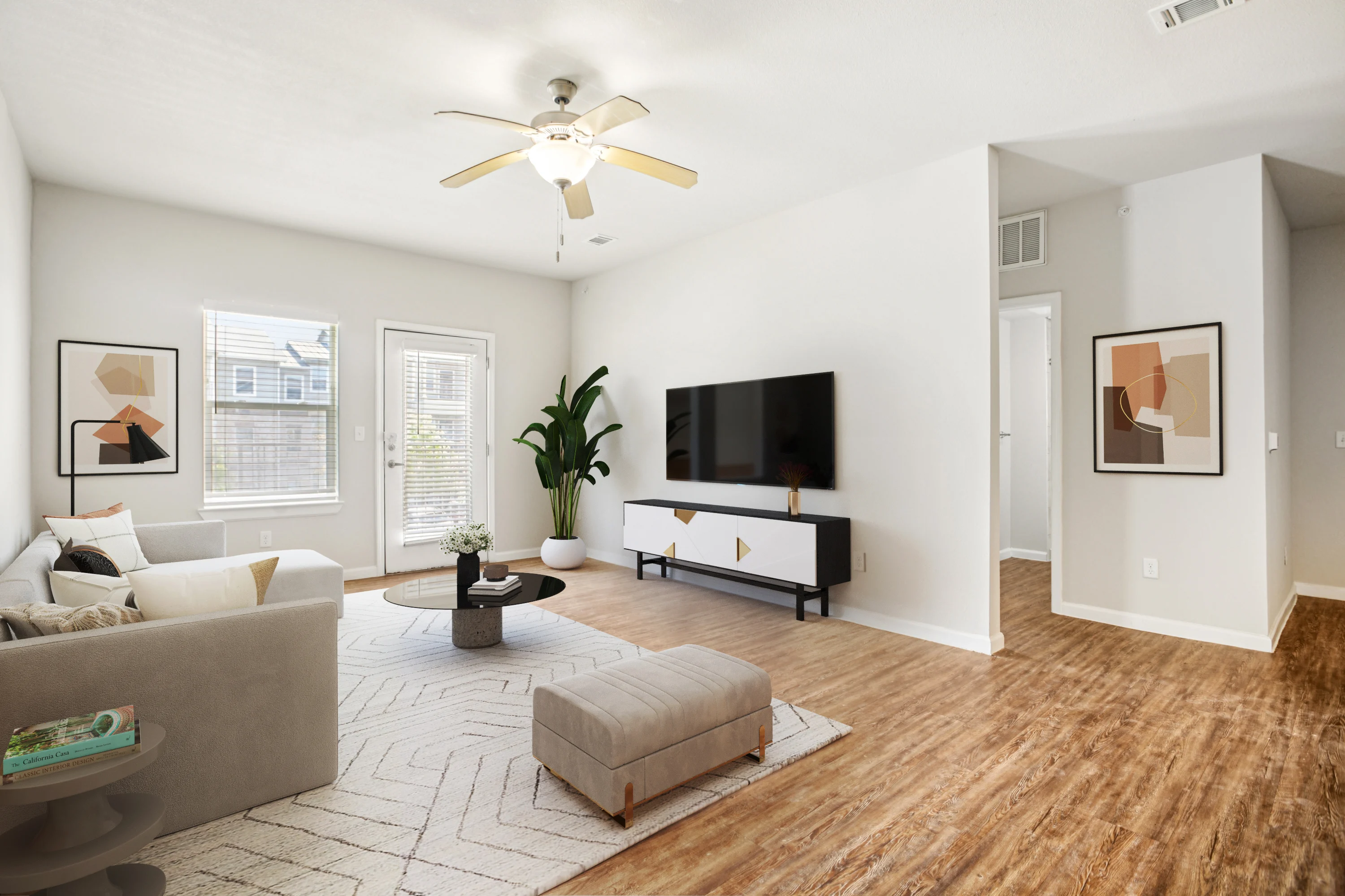 A modern living room featuring a gray couch, a round coffee table, and a white media console with a flat-screen TV. There are large windows letting in natural light and a potted plant in the corner. The floor is wooden and there is a patterned rug.
