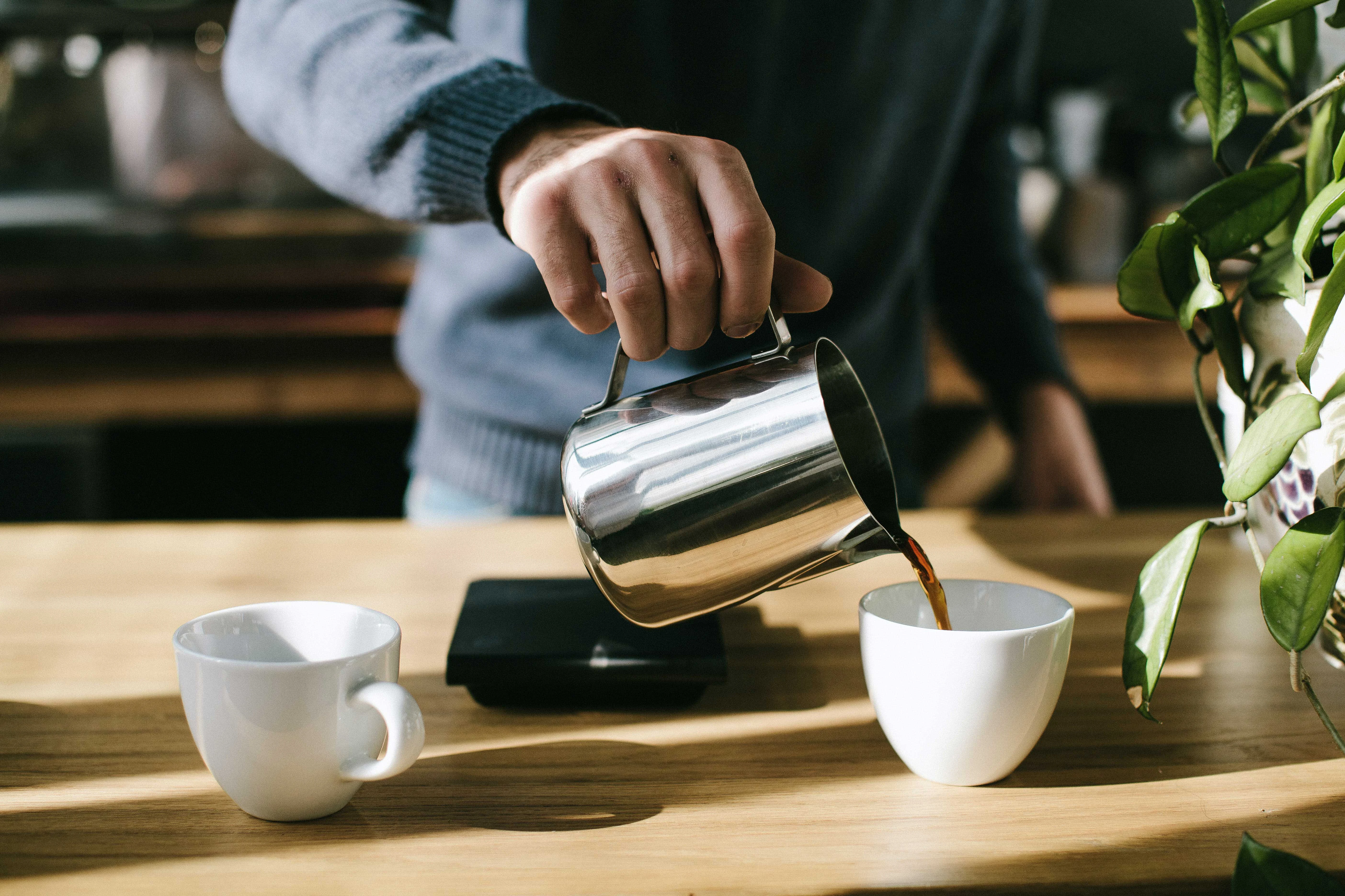 A person pouring coffee from a metal pitcher into a white cup, with another cup nearby, on a wooden table. There are elements of a kitchen environment in the background.