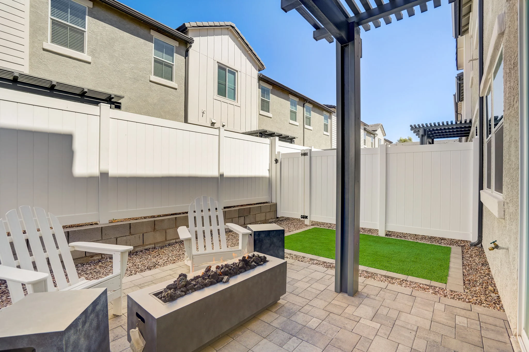 A cozy outdoor patio featuring two white adirondack chairs, a square fire pit with rocks, artificial grass, and a fenced enclosure.