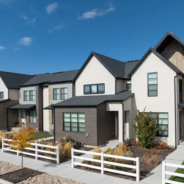A row of modern houses featuring a combination of brick and white siding with triangular roofs and large windows. A paved walkway leads to the entrances, surrounded by well-maintained landscaping and decorative fences.