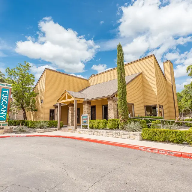 A modern yellow building with a sloped roof and large windows, surrounded by greenery. A sign reads 'Tuscany'.