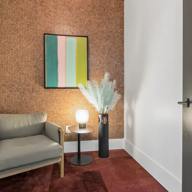 Stylish Relaxation Corner A cozy corner featuring a light gray armchair, a small black side table with a round lamp, a tall vase with decorative greenery, and abstract art on the wall.