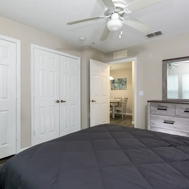 A cozy bedroom featuring a bed with a dark quilt, a ceiling fan, and light-colored walls. There are two open closet doors on the left and a dresser on the right, with a mirror above it. In the background, a doorway leads to another room with a dining area.