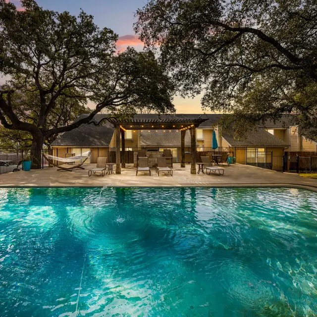 An inviting swimming pool area with crystal clear water surrounded by trees at dusk. The setting features a patio with seating and soft lighting.