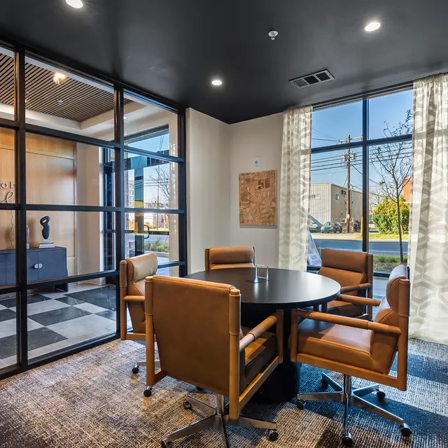 A modern meeting room featuring a black round table surrounded by four brown leather chairs, with large windows letting in natural light and a glass partition separating the space from an entryway with a wooden wall. The decor includes a large mounted TV and patterned flooring.