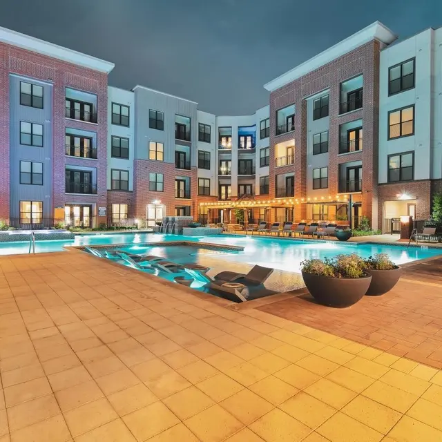 Modern Apartment Complex with Pool Area A modern apartment complex with a pool area at dusk. The scene shows a well-lit building with multiple floors and balconies, surrounding a spacious pool with lounge chairs.