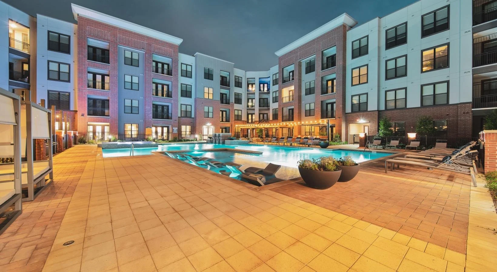 A modern apartment complex with a pool area at dusk. The scene shows a well-lit building with multiple floors and balconies, surrounding a spacious pool with lounge chairs.