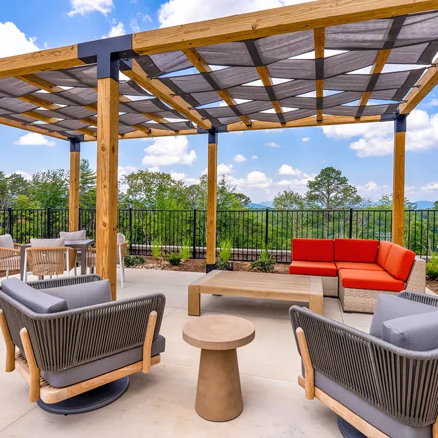 A modern rooftop terrace featuring a wooden pergola with shaded seating areas. The space has a stylish combination of gray and orange furniture, including two gray lounge chairs, a red couch, a small round table, and a backdrop of lush greenery and blue skies.