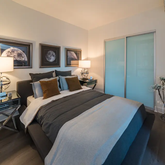 A modern bedroom featuring a bed with gray and white bedding, decorative pillows, two bedside lamps, and artwork on the walls.