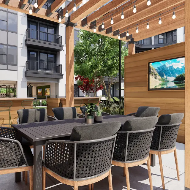A spacious outdoor dining area with large table and comfortable chairs, surrounded by wooden slats and greenery. A television mounted on the wall is visible in the background.