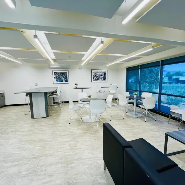 A modern office break room with tables and chairs, featuring gray cabinetry, a microwave, and large windows letting in natural light.