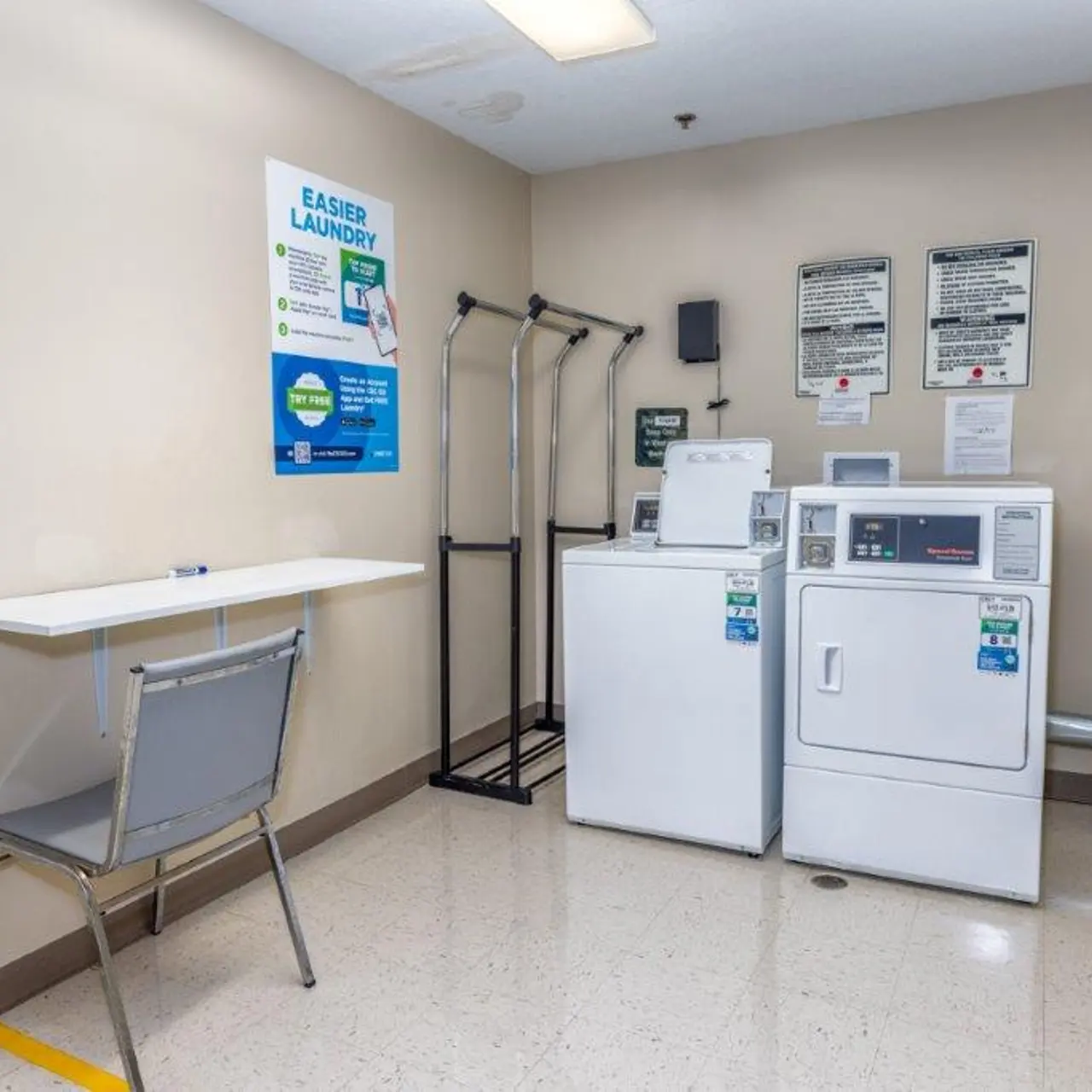 A small laundry room featuring a washing machine and dryer alongside a folding table and a chair. The walls are painted in a light color with various instructional signs displayed.