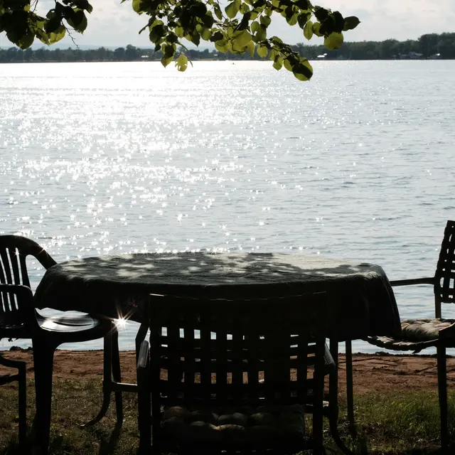 Barnacle Buds A table and chairs set beside a calm lake with sparkling water in the background.