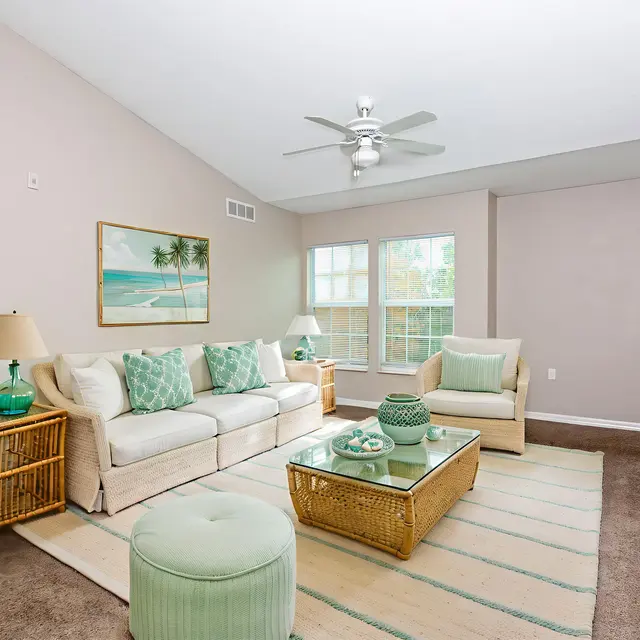 A bright and airy living room featuring a light color scheme with cream-colored furniture, green accents, and large windows allowing natural light. Decor includes a coffee table with decorative items, a comfortable chair, and a rug with subtle stripes.