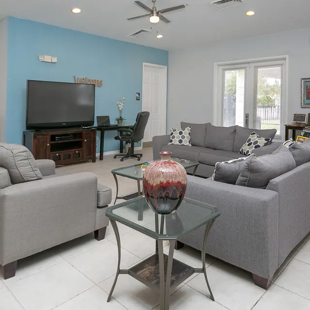 A modern living room with a grey sectional sofa, a glass coffee table with a decorative vase, and a large TV on the wall. There are light-colored tiles and walls, and an office chair can be seen in the background.