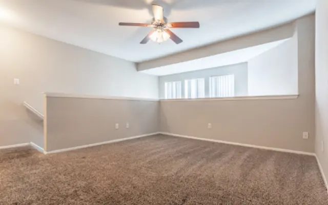 Spacious Living Room A spacious living room with carpeting, a ceiling fan, and large windows allowing natural light. The walls are painted in a light gray color.