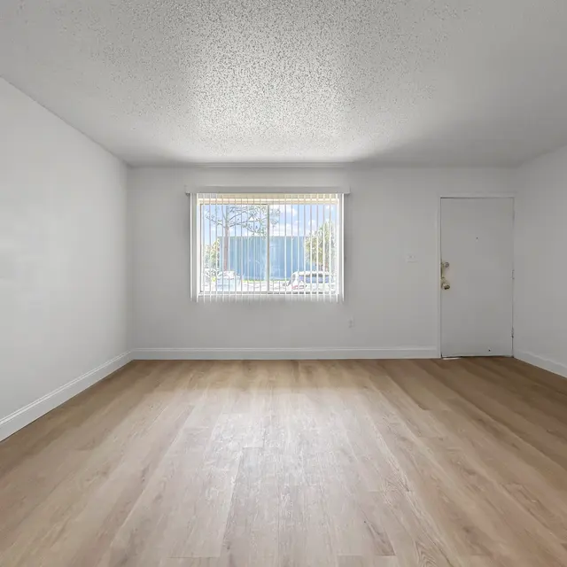 Spacious Unfurnished Room An empty room with light wooden flooring and white walls. A window with blinds is visible on one wall, and a door is present on the right side.