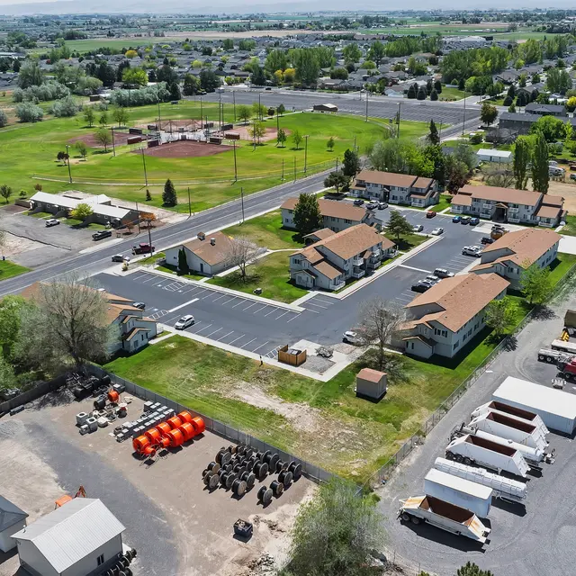Aerial Neighborhood Overview Aerial view of a residential neighborhood with several houses, a park, and open fields. The area includes a baseball diamond and various parking spaces with equipment and containers.
