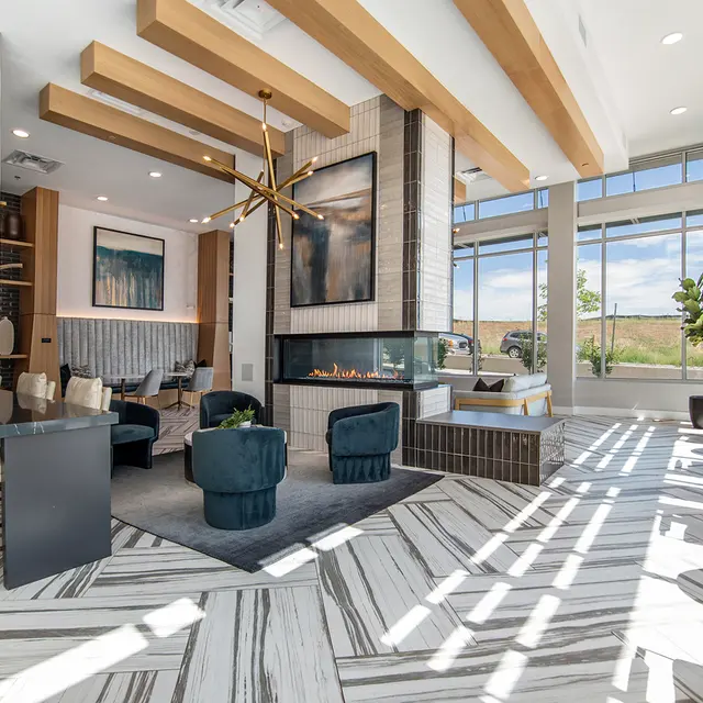 A stylish living area with large windows letting in natural light. The room features a modern fireplace, comfortable seating, and decorative elements like a statement chandelier. The floor has a distinct striped pattern, and there are plants visible outside through the glass.