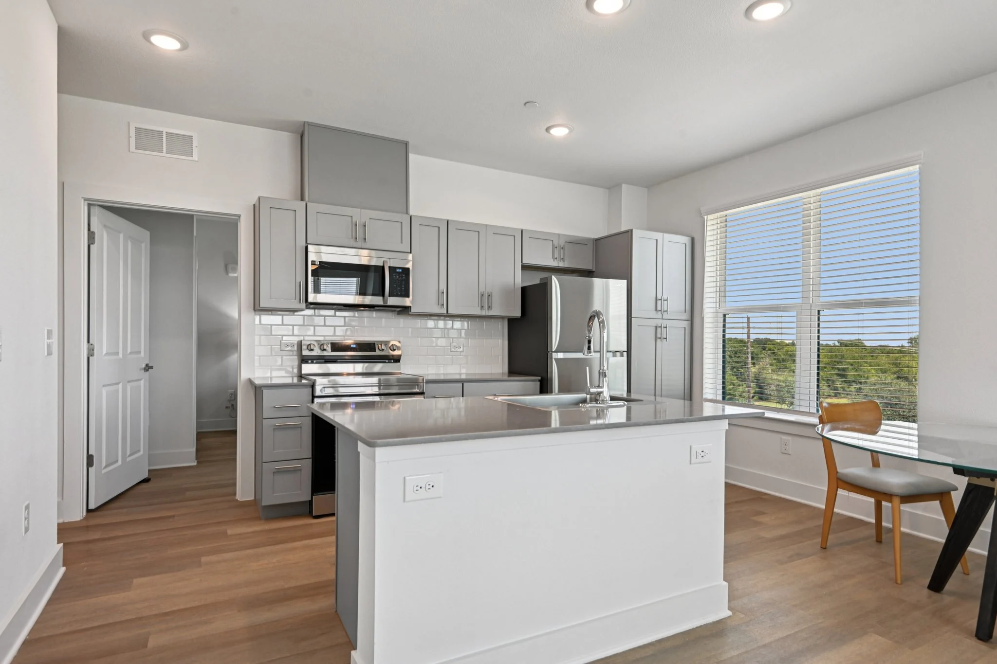 A modern kitchen featuring gray cabinetry, stainless steel appliances, and a large island with a countertop. The kitchen has ample natural light from large windows.