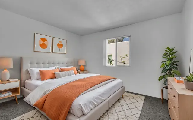 A modern bedroom featuring a large plush bed with orange and white bedding, two bedside lamps, and a wooden dresser. The room has a large window with a view of greenery outside and is decorated with minimalistic art on the walls.
