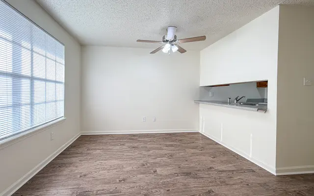 Empty living room with wooden floor and ceiling fan, featuring a window with blinds on one side and an open kitchen area.