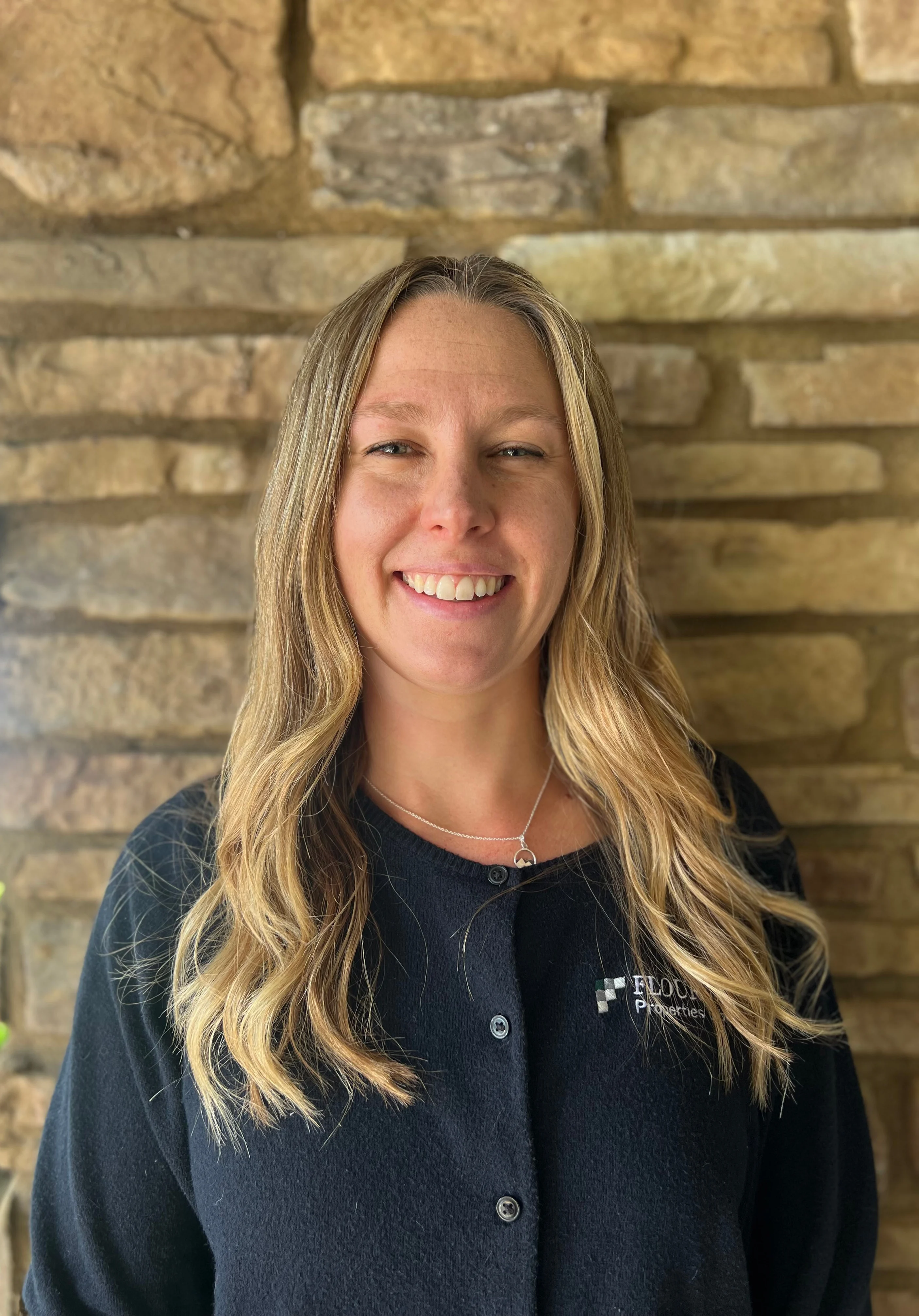 Smiling Woman in Front of Stone Wall A smiling woman with long hair standing in front of a stone wall, wearing a black cardigan.
