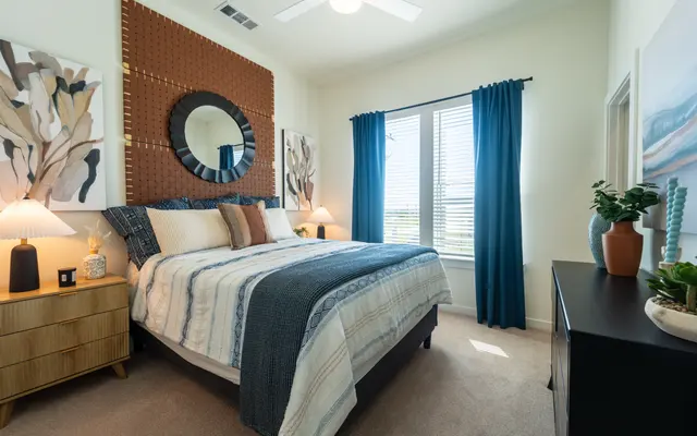 A cozy modern bedroom featuring a large bed with a patterned blue and white blanket. There are decorative pillows on the bed and a brown accent wall behind the headboard. Two bedside lamps illuminate the space, and there are window treatments showcasing light streaming in. The room has a neutral carpet and various decorative artwork on the walls.