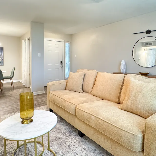 A cozy living room featuring a beige sofa with pillows, a round coffee table with a yellow vase, and a dining area in the background with a glass table and modern chairs.