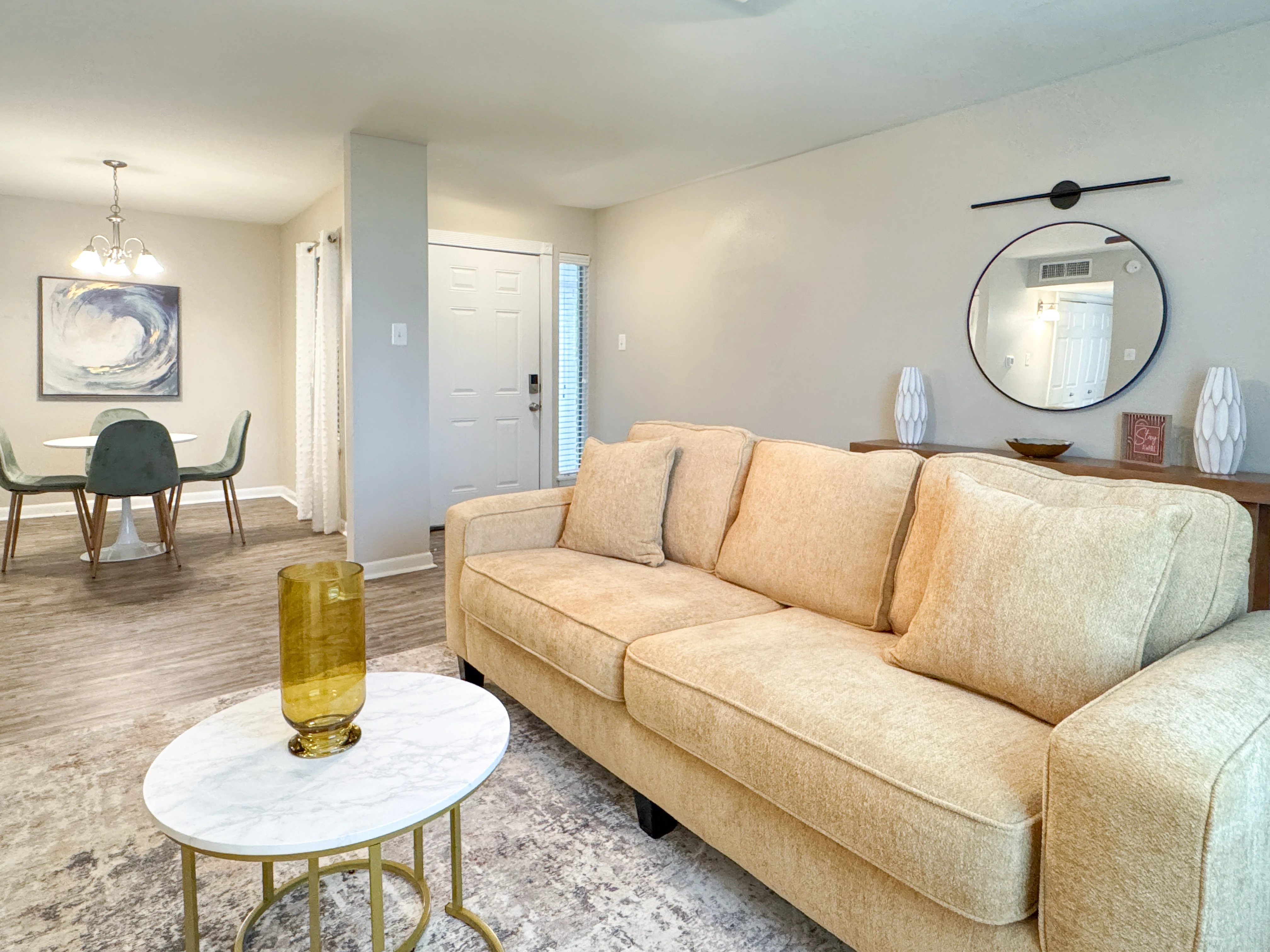 A cozy living room featuring a beige sofa with pillows, a round coffee table with a yellow vase, and a dining area in the background with a glass table and modern chairs.