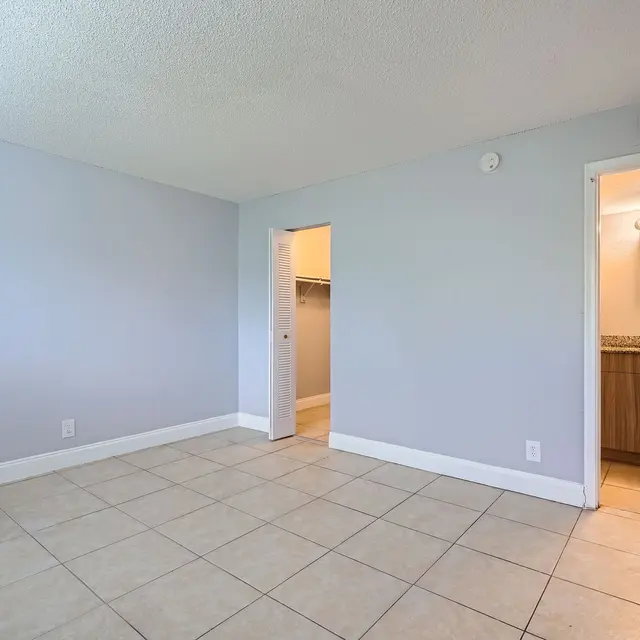 A spacious empty room with light blue walls and tile flooring. A doorway leads to a bathroom visible on the right, while another doorway leads to a closet area on the left.