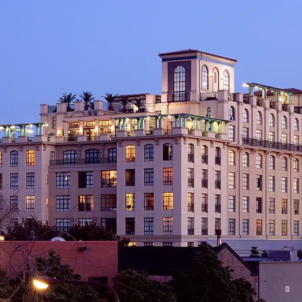 A historic hotel building at dusk with multiple stories and large windows, featuring a rooftop terrace adorned with palm trees.