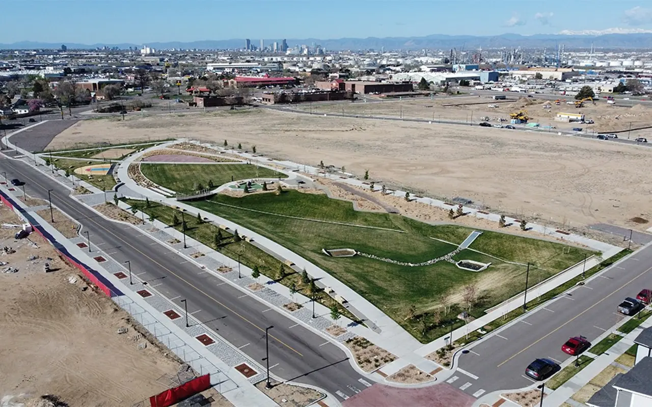 Aerial view of a newly designed park with green grass and pathways, set in an urban area, with a city skyline in the background.