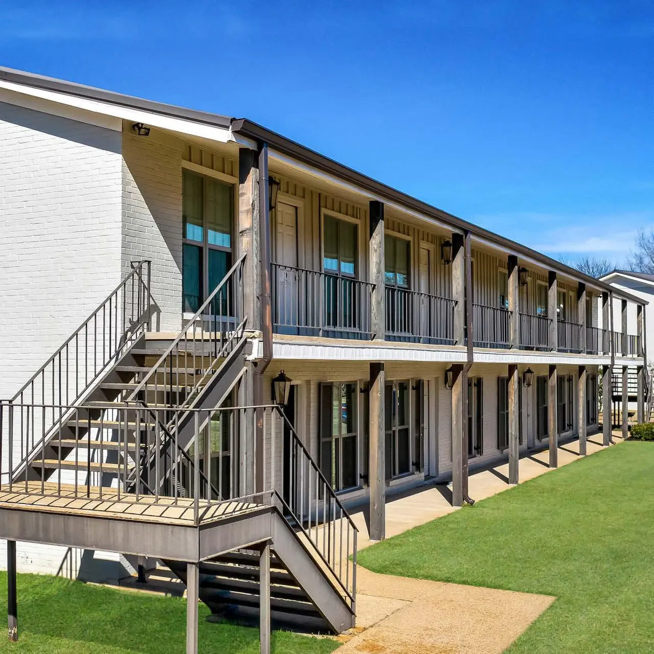 A modern two-story apartment building with balconies and stairs, surrounded by green grass under a clear blue sky.