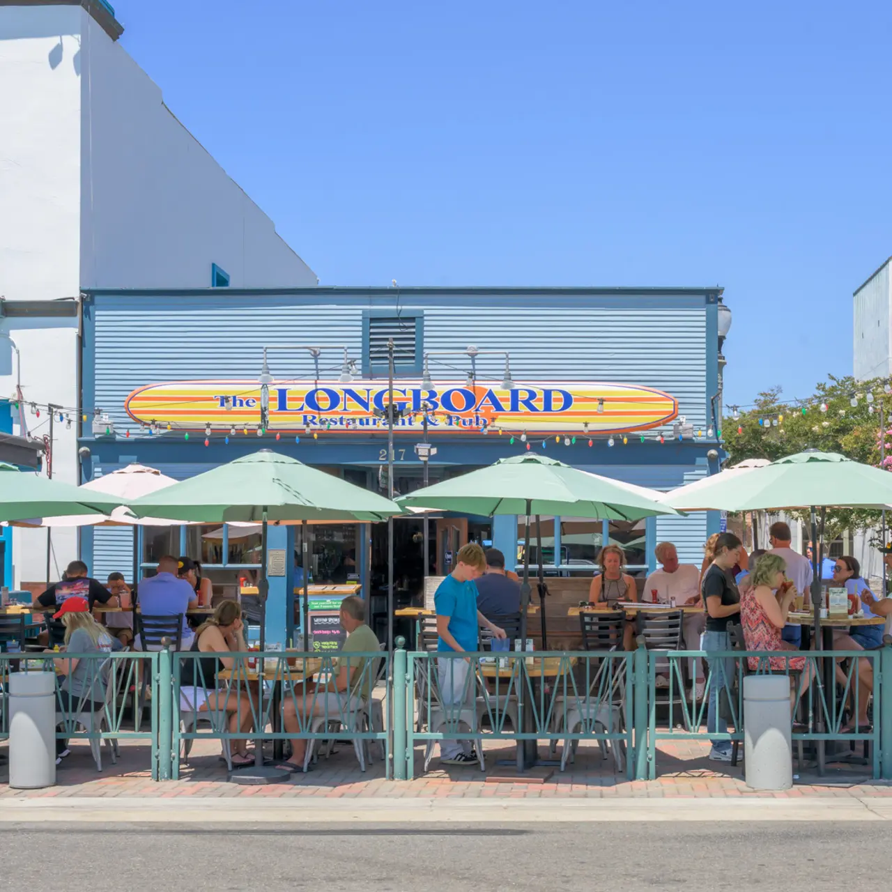 Outdoor dining area at The Longboard restaurant with people seated under green umbrellas.