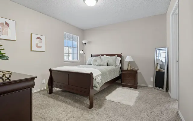 A cozy bedroom featuring a wooden bed with light-colored bedding, a bedside table with a lamp, and a full-length mirror. Soft carpeting and minimal decor enhance the inviting atmosphere.