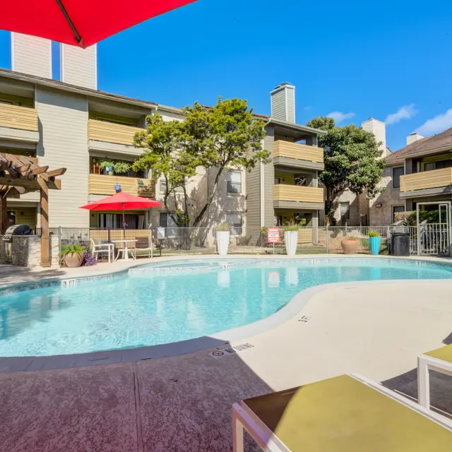 A serene apartment complex pool area featuring a turquoise swimming pool surrounded by lounge chairs. Red umbrellas provide shade, and there are trees and plants around. Two stories of apartment buildings are visible in the background under a clear blue sky.