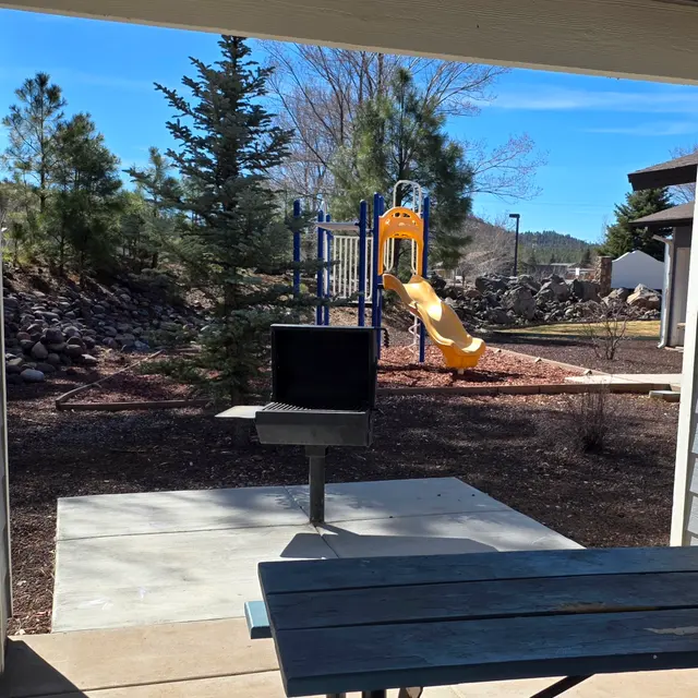 A view from a covered area showing a picnic table and a grill, overlooking a playground with a yellow slide and climbing structure. There are trees in the background and a clear blue sky.